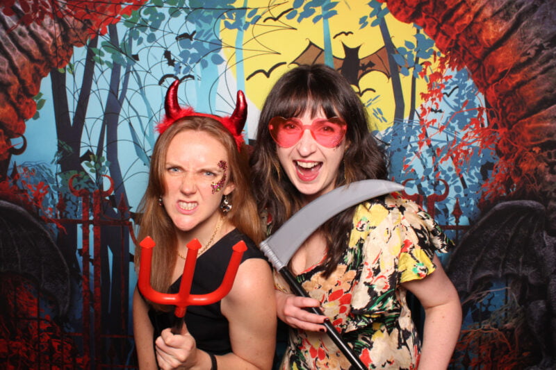two women are pointing at the camera with devil head hair ornaments on their heads halloween party photo booth 2, in the style of hellish background